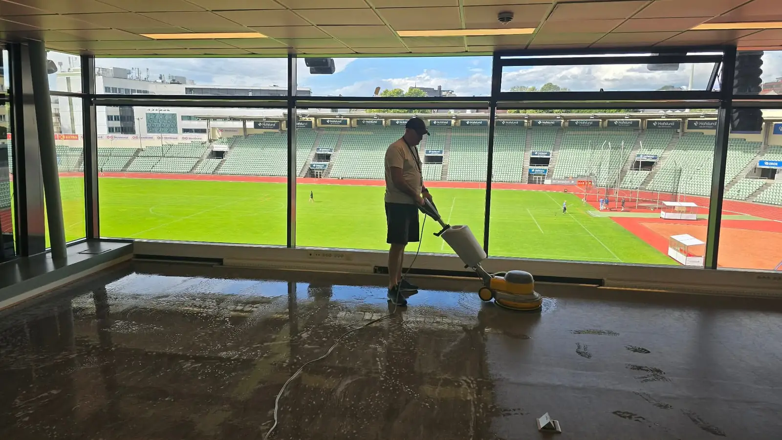 Renholder fra Finstad Renhold maskinskurer gulvet i en VIP-losje på Bislett stadion i Oslo, med løpebanen og gressmatta synlig gjennom vindusveggen